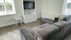 A modern living room with a grey sectional sofa, a white entertainment unit with a TV, and a small white side table. - Studio Apartment in Londonderry (Londonderry)