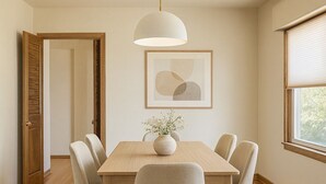 A dining area with a wooden table, six chairs, a vase with flowers, a pendant light, and a framed abstract artwork on the wall.
