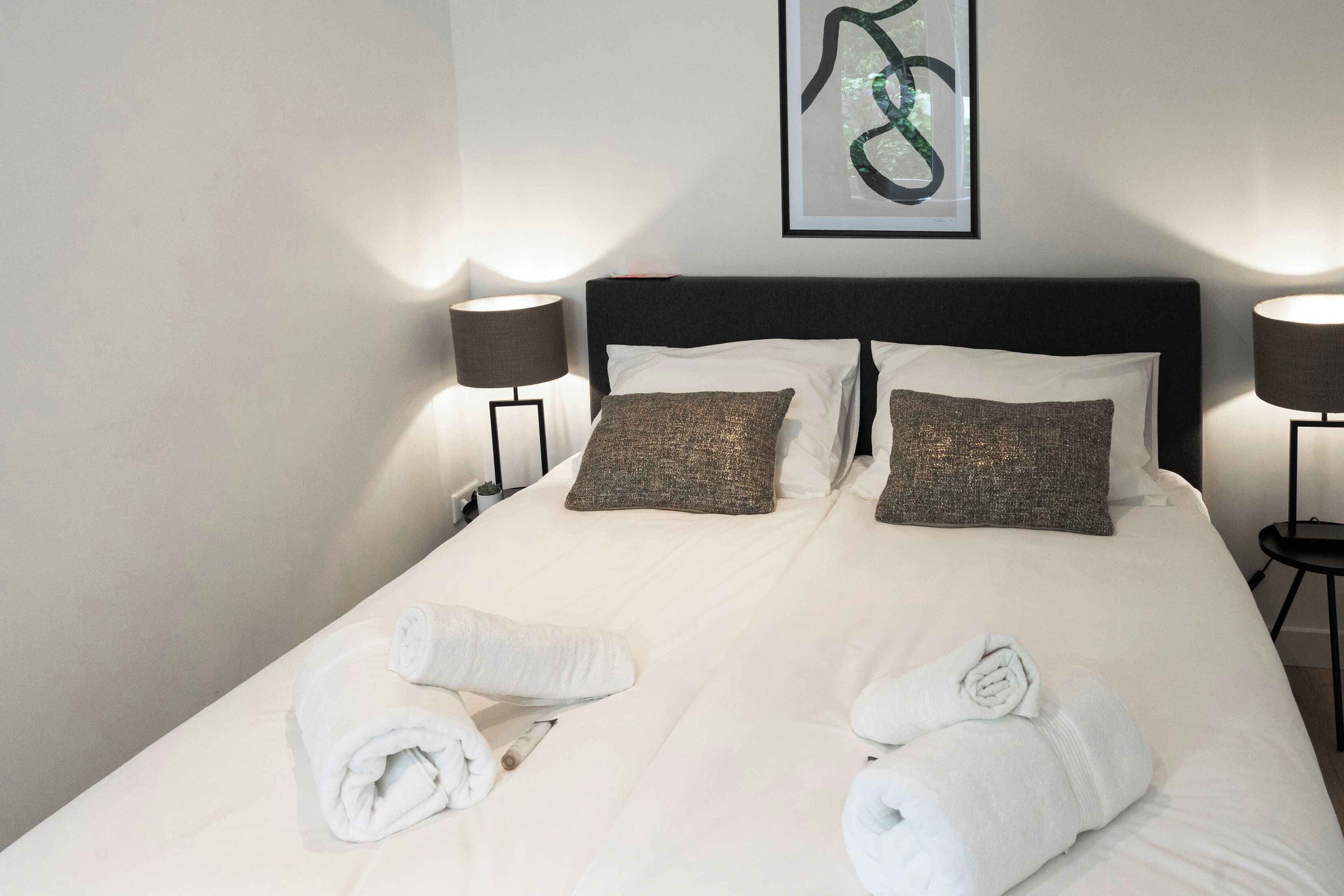 Een netjes opgemaakt bed met witte lakens en twee opgerolde handdoeken, een zwart hoofdeinde en een lijst met abstracte kunst aan de muur.