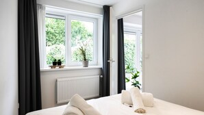 A hotel room with a bed, a window with curtains, a radiator, and a view of greenery outside.