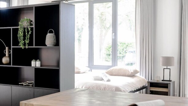 A modern bedroom with a large bed, a bedside table, and a nightstand. There is a window with curtains, a bookshelf, and a desk with chairs.
