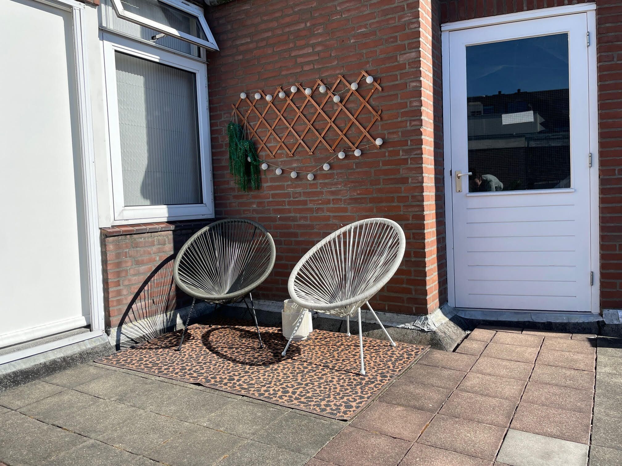 Un bâtiment en briques avec deux chaises en osier blanches posées sur un tapis à motifs, une porte blanche et une fenêtre avec un moustiquaire.
