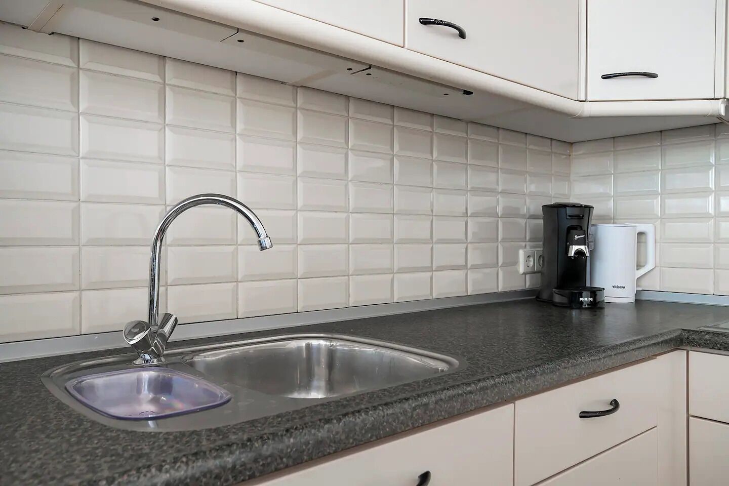 A kitchen with a double sink, white cabinets, a coffee maker, and a kettle.