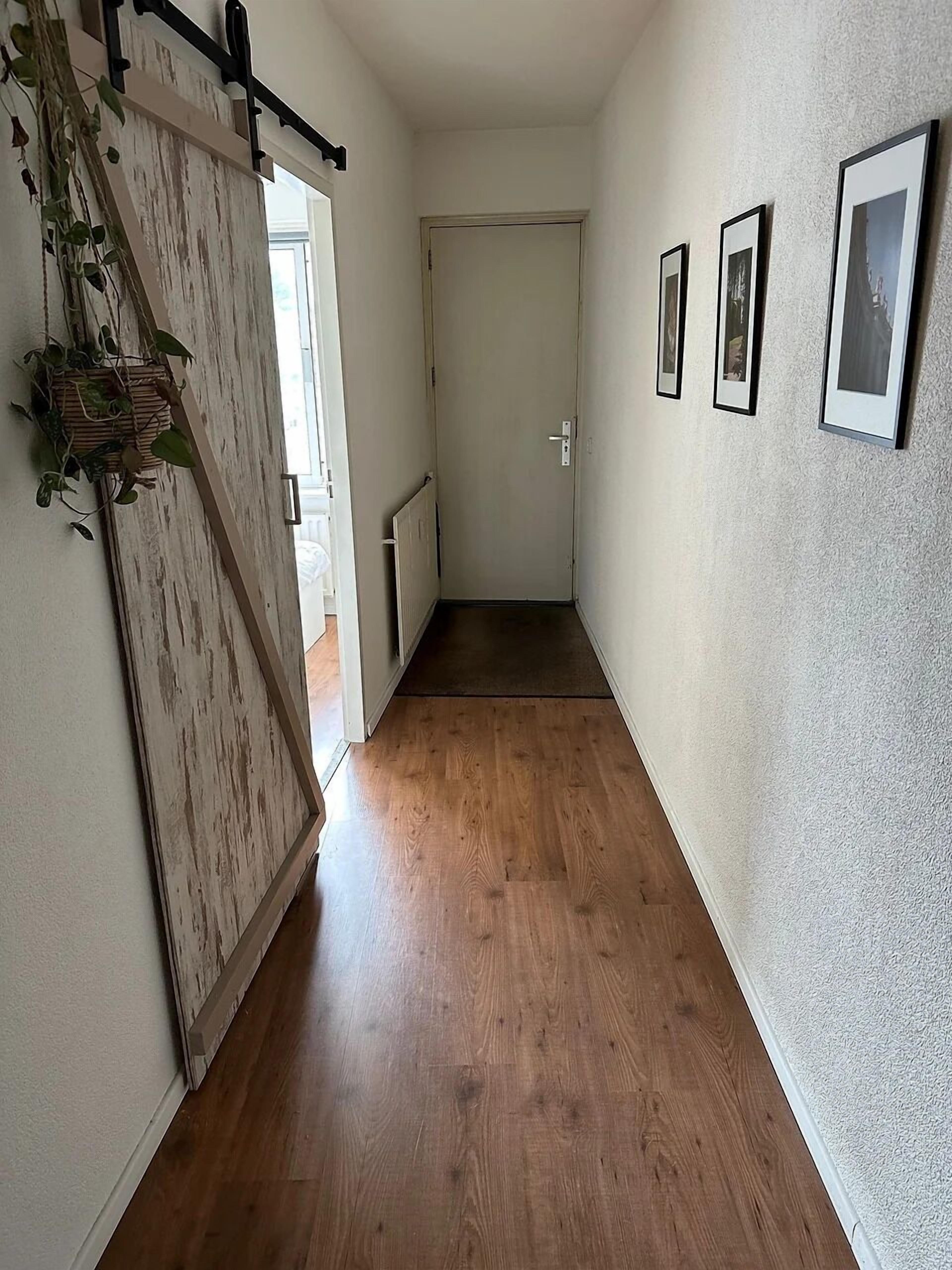 A hallway with wooden flooring, a white door at the end, and three framed pictures on the wall.