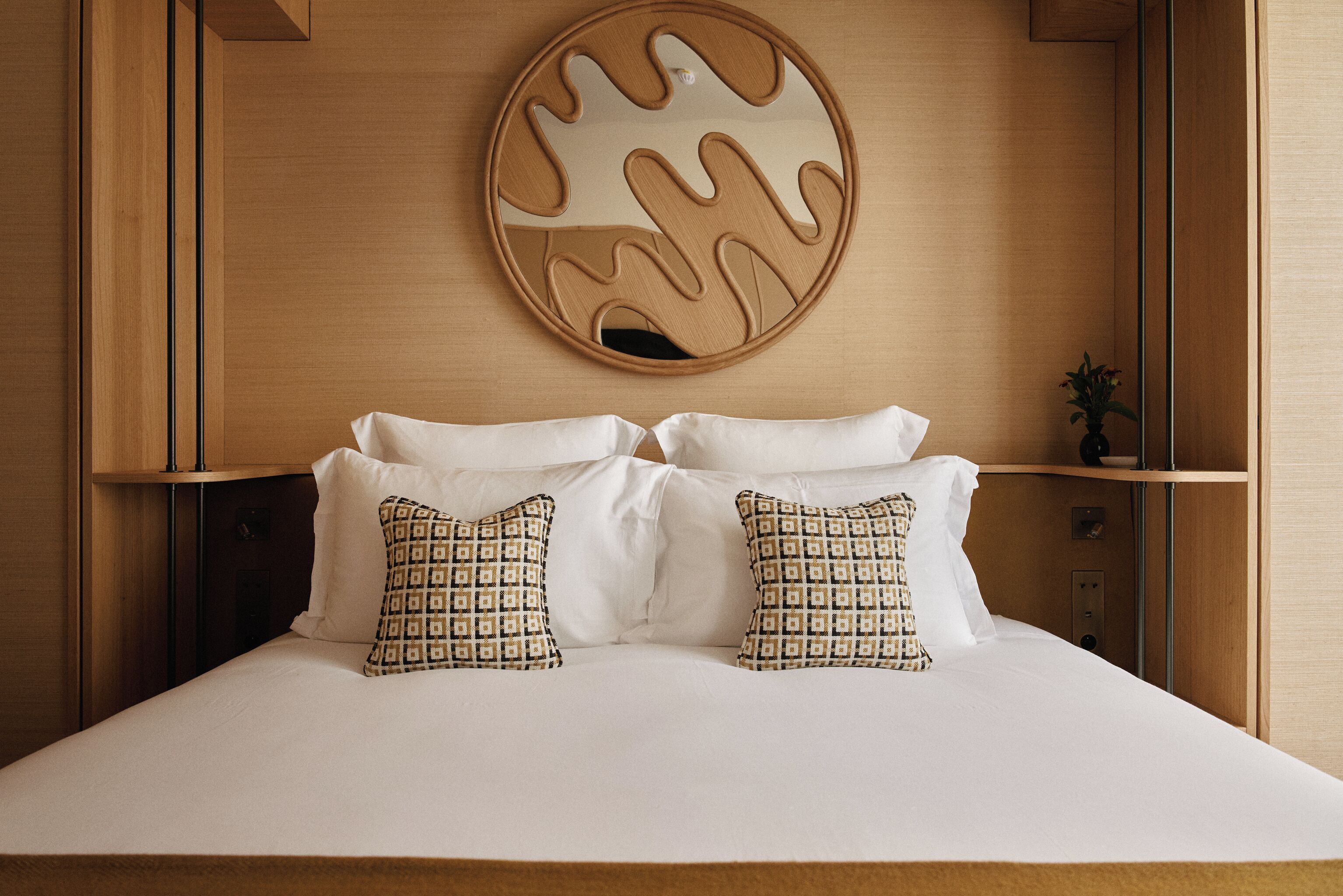 Una cama bien tendida con almohadas blancas y estampadas, un cabecero de madera y un espejo circular fijado a la pared con un diseño único.