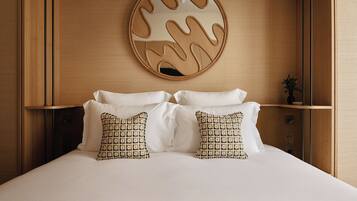 Una cama bien tendida con almohadas blancas y estampadas, un cabecero de madera y un espejo circular fijado a la pared con un diseño único.