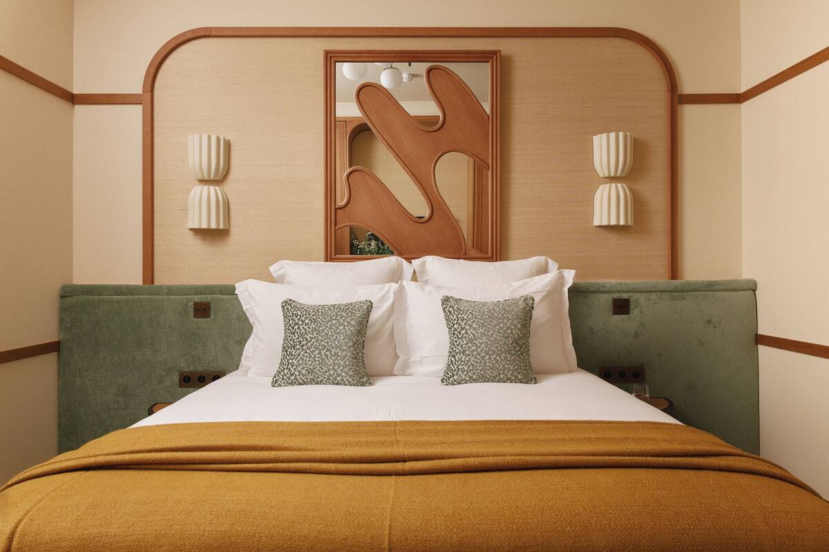 A hotel room with a bed, green upholstered headboard, white bedding, and a unique wall-mounted mirror.