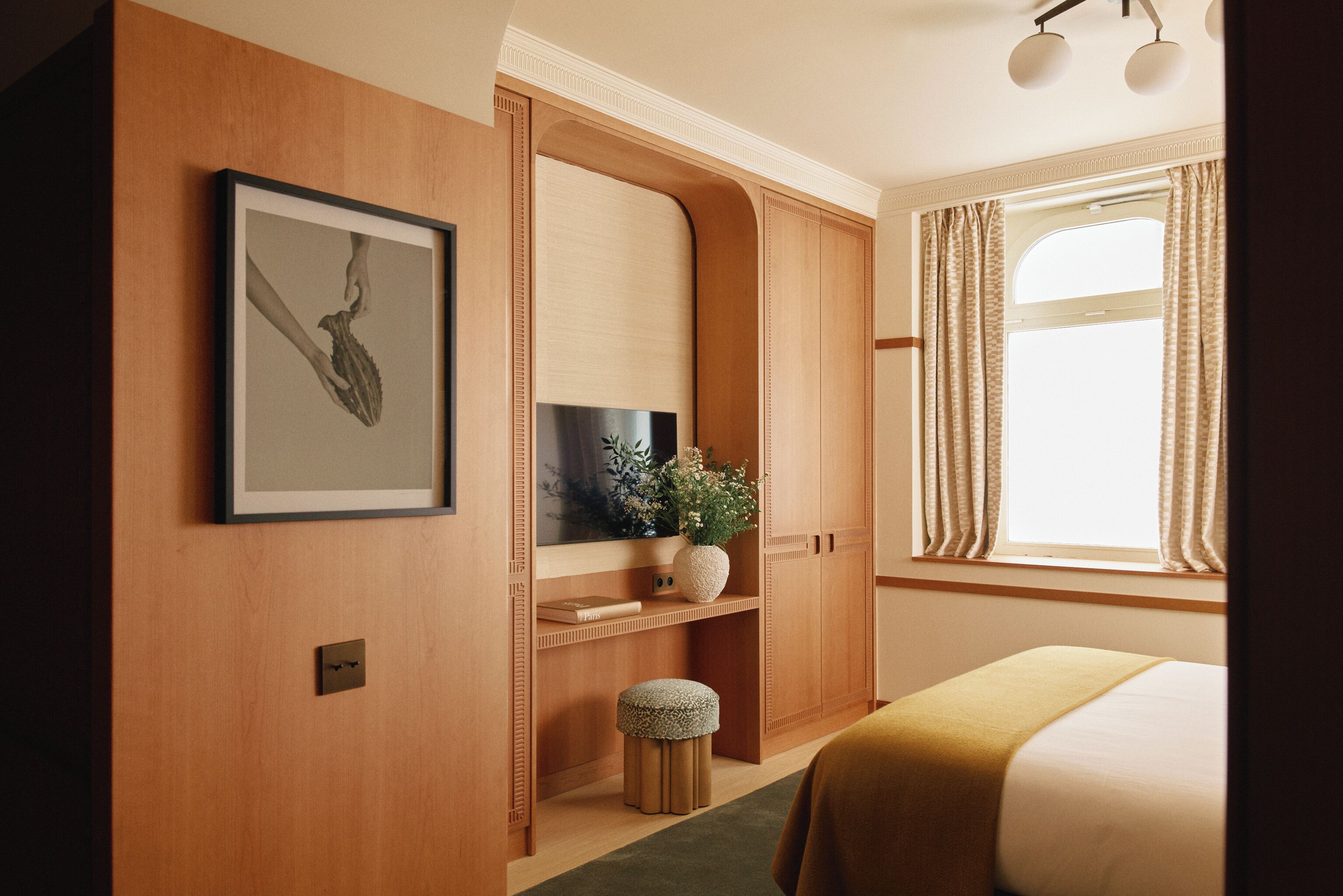 A hotel room with a wooden wardrobe, a framed picture, a potted plant, and a bed with a green rug.