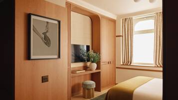 A hotel room with a wooden wardrobe, a framed picture, a potted plant, and a bed with a green rug.