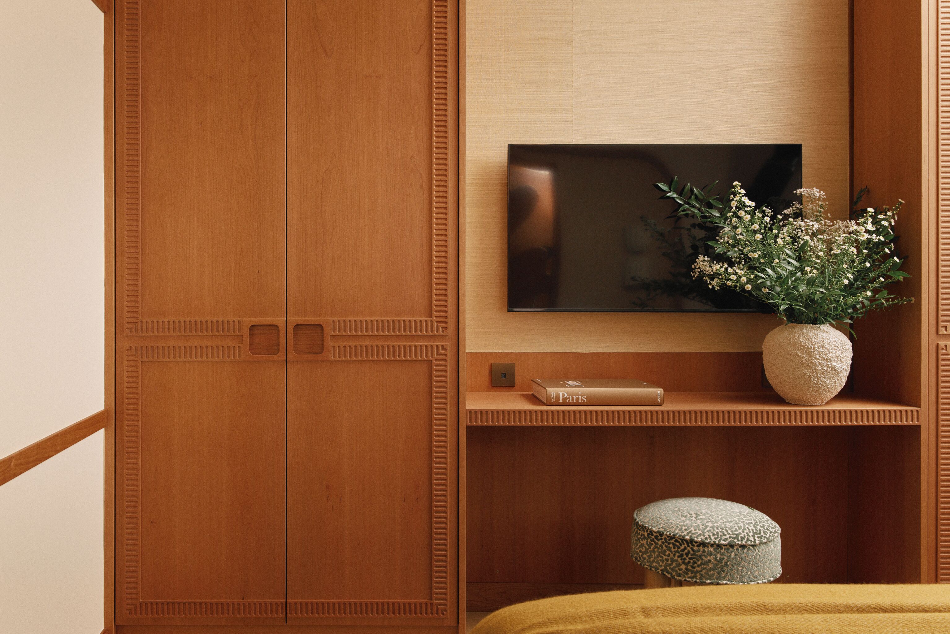 A wooden wardrobe with a flat-screen TV mounted above it, a vase with flowers, and a book titled "Paris" on a shelf.