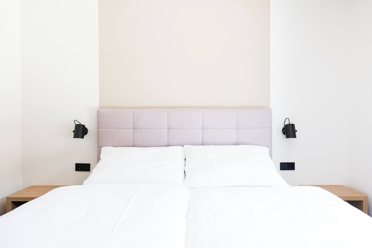 Un lit bien fait, avec du linge de lit blanc, une tête de lit de couleur claire et deux lampes de chevet noires, une de chaque côté.