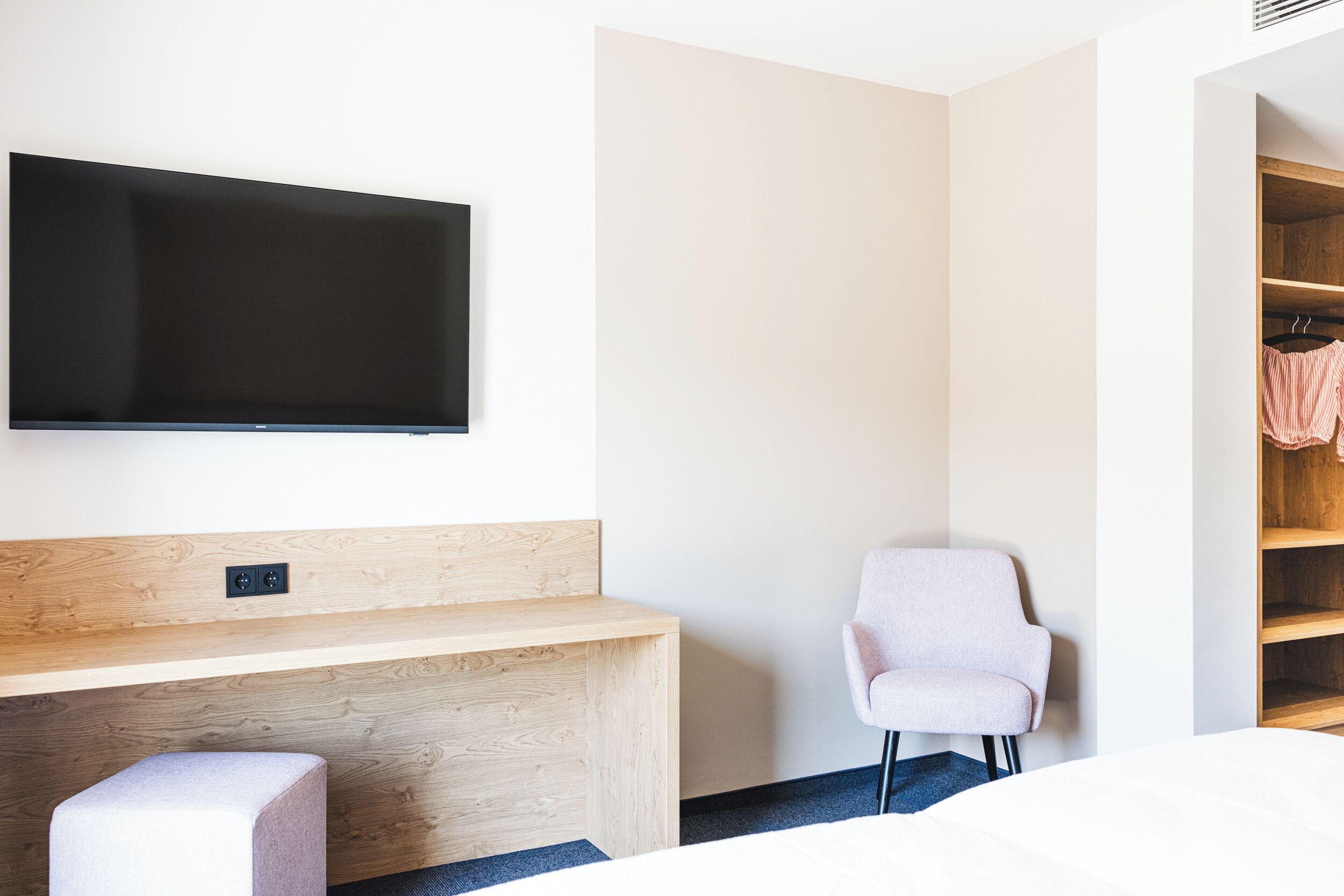 Une chambre d’hôtel moderne dotée d’un téléviseur à écran plat fixé au mur, d’un bureau en bois, d’un fauteuil confortable et d’une armoire intégrée avec espace pour suspendre les vêtements.