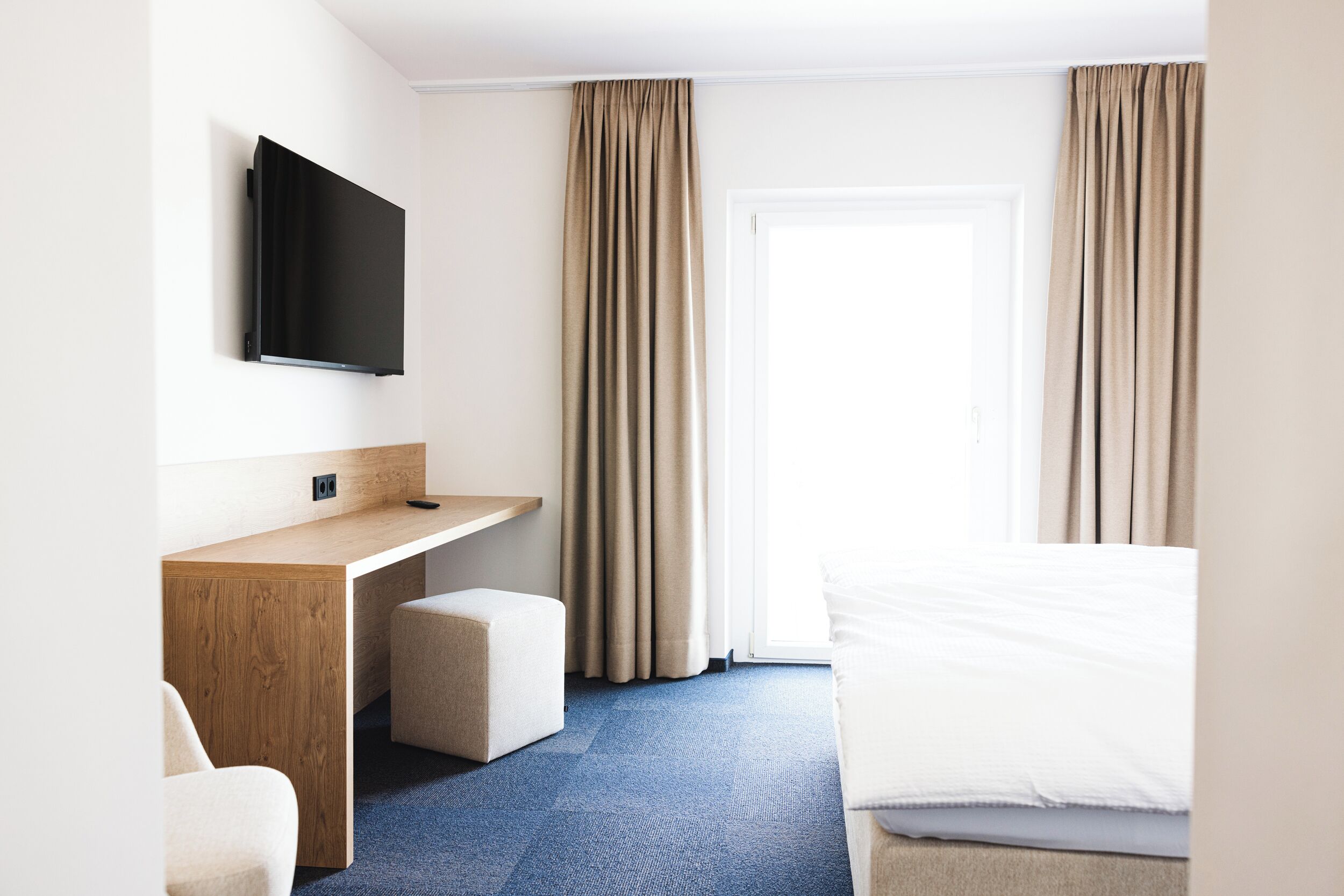 Hotelkamer met een wandgemonteerde flatscreen-tv, een houten bureau, een klein voetenbankje, een bed met wit beddengoed en een groot raam met beige gordijnen.