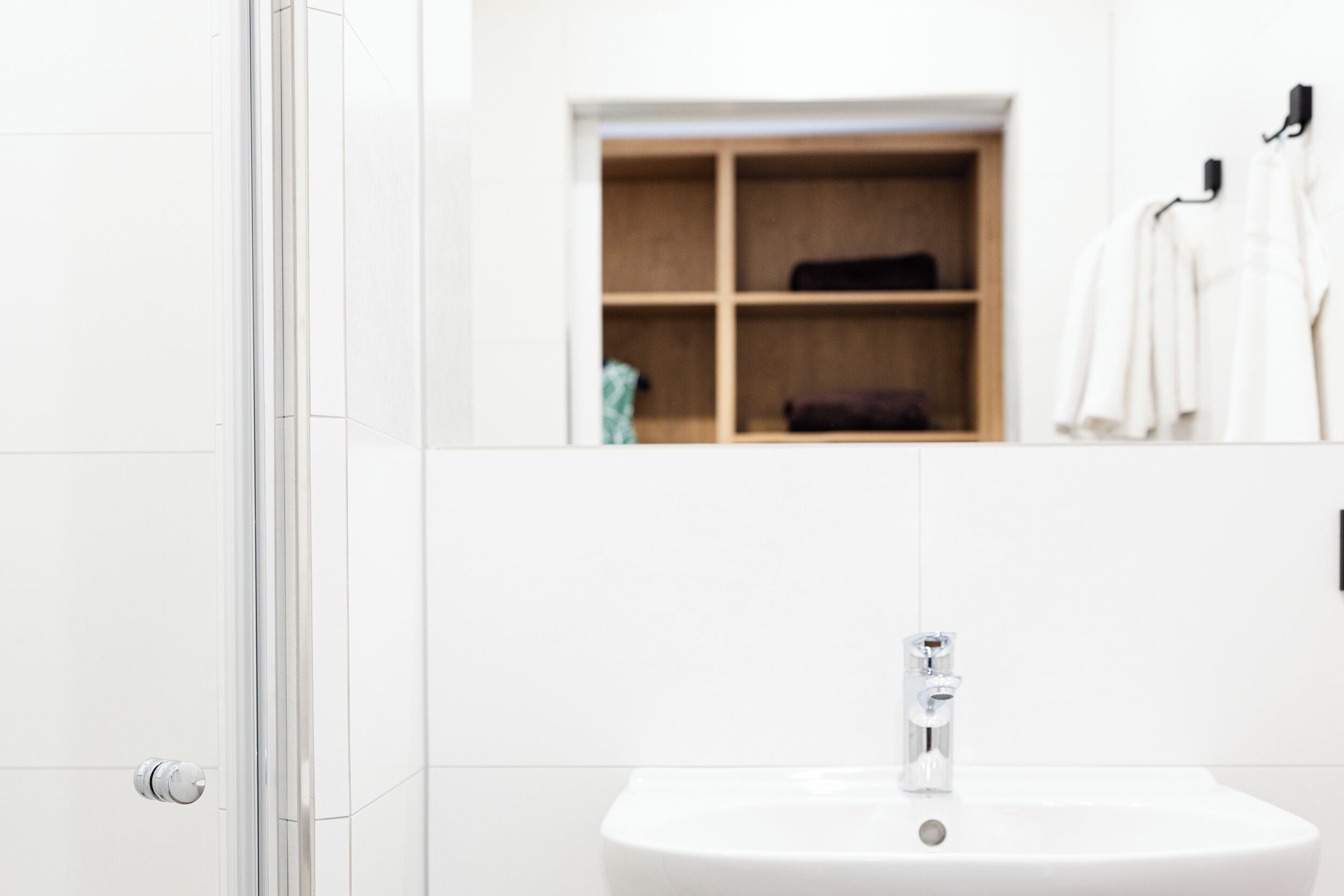 A bathroom with a glass shower door, a white sink with a chrome faucet, and a wooden shelf with towels and a robe.