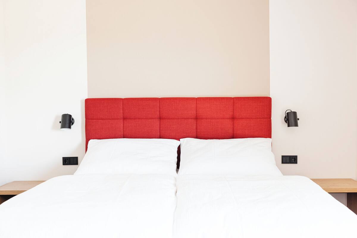 Un lit avec du linge de lit blanc, une tête de lit rembourrée en tissu rouge et deux lampes de chevet.