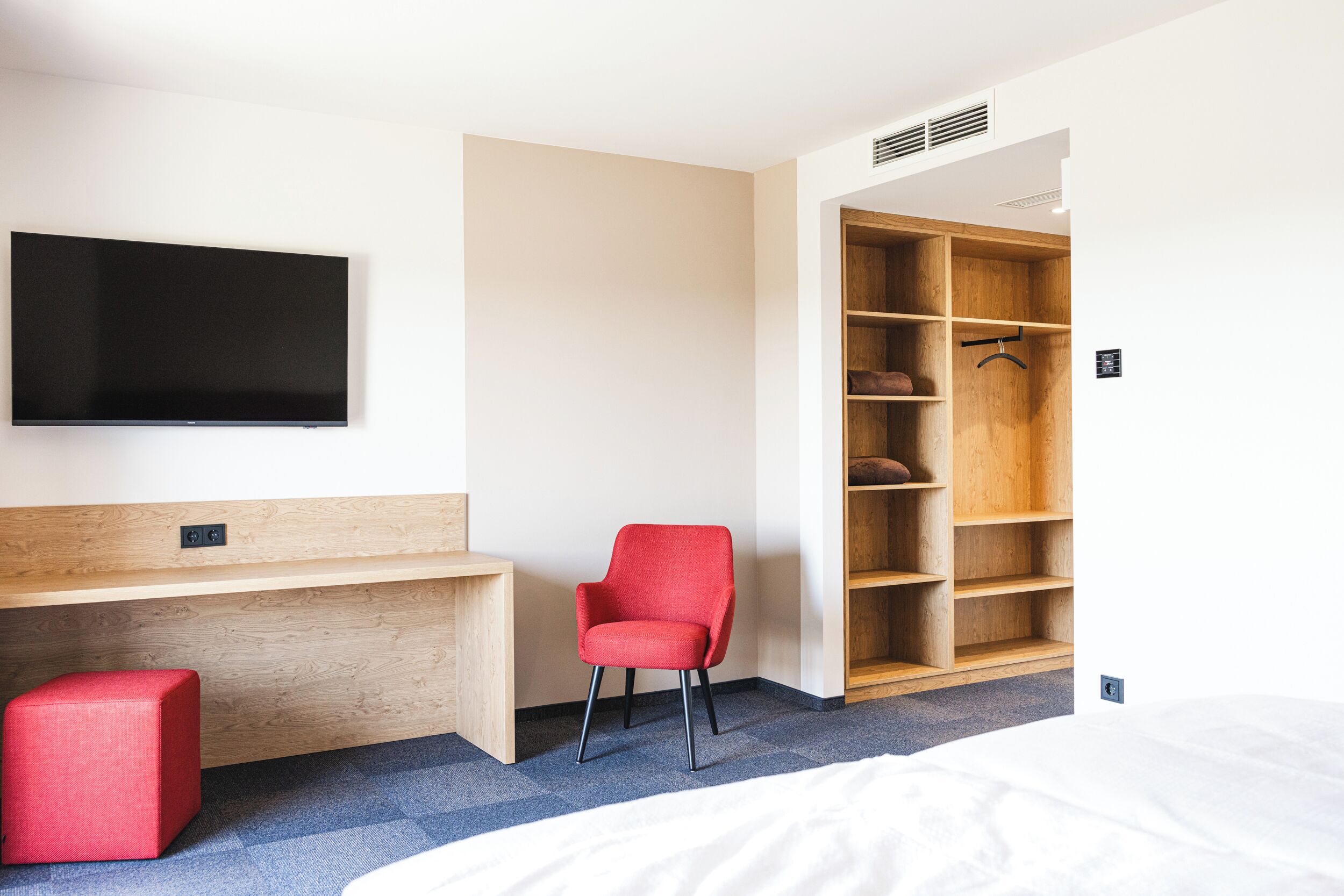 Ein Hotelzimmer mit einem Bett, einem roten Sessel, einem Holztisch, einem an der Wand montierten Fernseher und einem begehbaren Kleiderschrank.