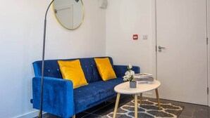 A room with a blue sofa, yellow cushions, a round table with a vase of flowers, and a black floor lamp. - Cozy Garden Studio in St Albans (St Albans)