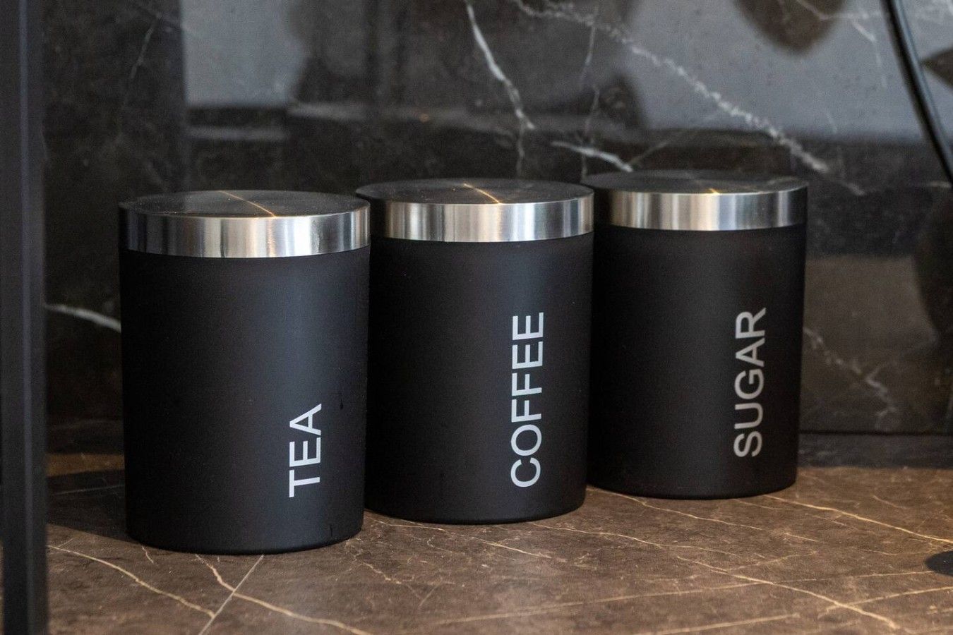 Three cylindrical containers labeled tea, coffee, and sugar on a marble surface.