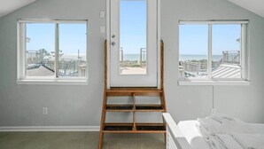 A room with a sloped ceiling, a wooden ladder leading to a door, and three windows offering views of a beach and buildings. - Coastal Aire - Gulf Front 3 bed, 4 bath! (Port St. Joe)
