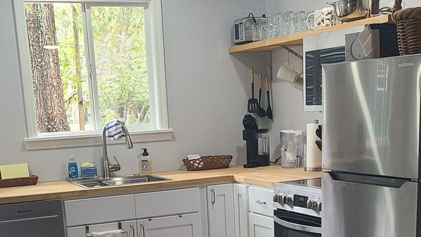 Una cocina moderna con armarios blancos, electrodomésticos de acero inoxidable, horno empotrado y ventana con vistas a árboles.