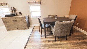 Zona pranzo con tavolo in legno, quattro sedie e un angolo cottura con pensile in legno e una finestra.