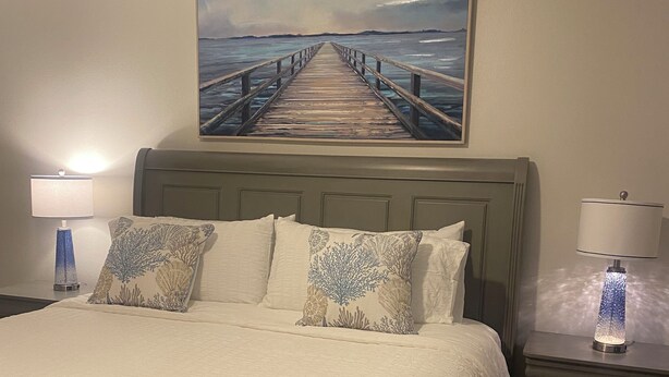 Una cama bien hecha con cabecera, dos mesitas de noche con lámparas, y un cuadro de un muelle en la pared.