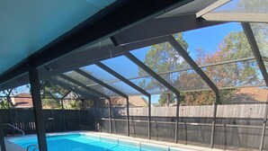 Una piscina exterior techada con superficie de agua azul cristalina, rodeada por una valla de madera y una estructura de techo metálico.