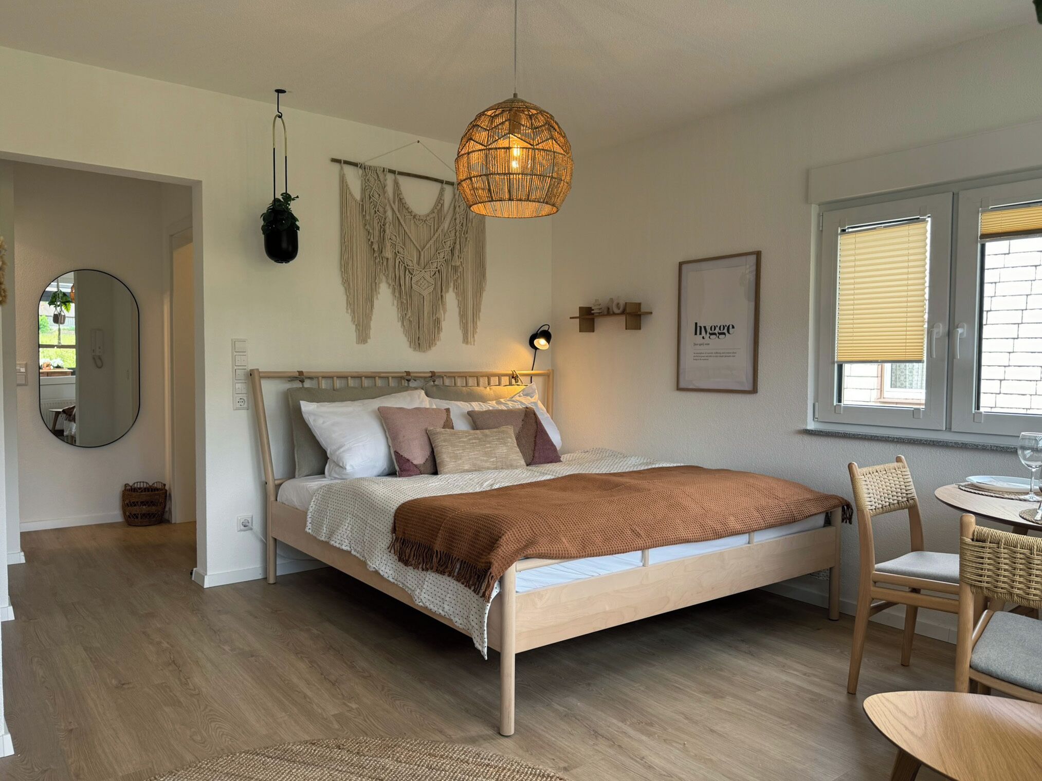 Une chambre avec un lit en bois, une décoration murale en macramé, un luminaire suspendu et une affiche encadrée sur le thème du « hygge ».