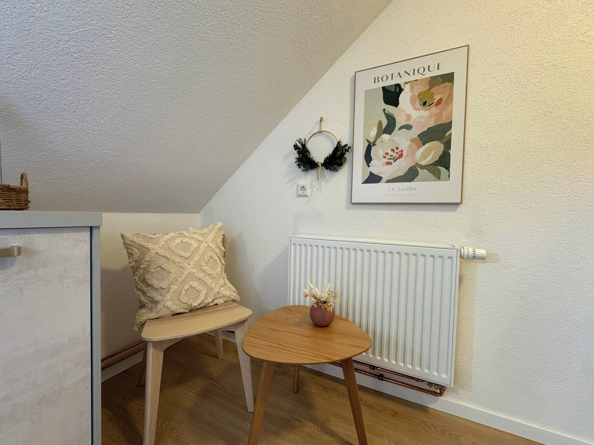 Una habitación pequeña y acogedora con un radiador blanco, una mesa de madera con un florero, un cojín decorativo e impresión botánica enmarcada en la pared.
