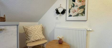 Una habitación pequeña y acogedora con un radiador blanco, una mesa de madera con un florero, un cojín decorativo e impresión botánica enmarcada en la pared.