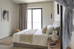 Une chambre moderne avec un grand lit, une table de chevet sur laquelle se trouvent un livre et un vase, une fenêtre avec des rideaux et une vue sur l’extérieur.