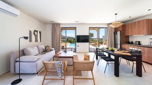 A modern living room with a sofa, coffee table, and chairs, a dining area with a table and chairs, and a kitchen with wooden cabinets and a large window.