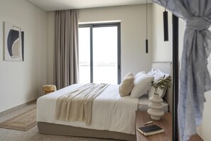 A modern bedroom with a large bed, a bedside table with a book and a vase, a window with curtains, and a view of the outdoors. - Massari Hills- Villa Nobu 4 (Rhodes)