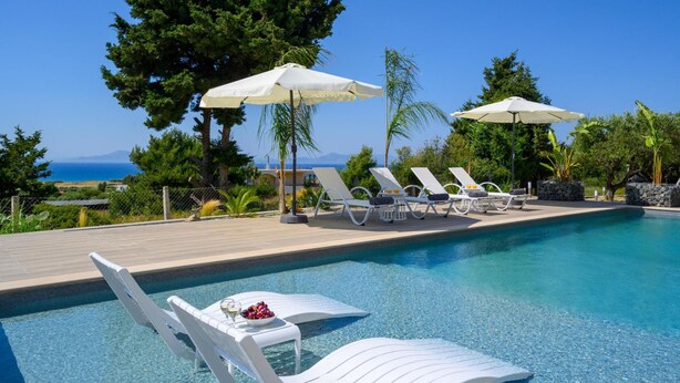 A poolside area with lounge chairs and umbrellas, a clear view of the sea, and a table with fruit and a glass. - Sunset View Paradisi Villa (Rhodes)