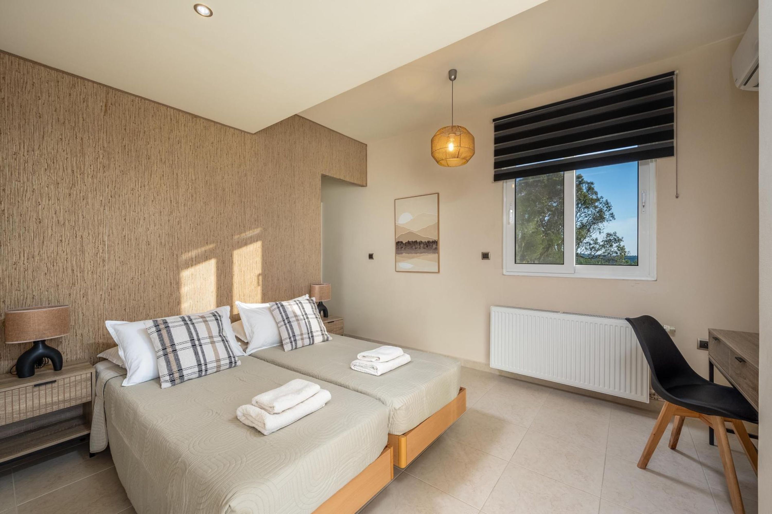 A modern bedroom with two beds, a radiator, a window with blinds, and a desk with a chair.
