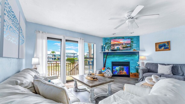 A spacious living room with a large window, a ceiling fan, a fireplace, and a TV. - 1-minute walk to beach, Townhome in Welcoming Old Florida Saint George Island! (Saint George Island)