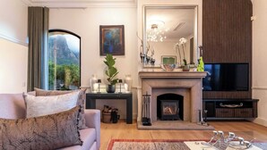 A living room with a sofa, a fireplace, a TV, and a view of a mountain through a window. - Stunning Views, Walk to Beach, Heated Splash Pool - Iparadesi (Cape Town)