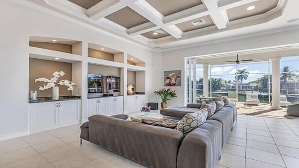 A spacious living room with a large gray sofa, a kitchen with white cabinetry, and a view of a pool and palm trees through large glass doors. - Tranquil Island Retreat with Modern Comforts (Marco Island)