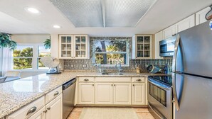 Une cuisine moderne dotée de plans de travail en granit, de meubles blancs, d’un réfrigérateur en acier inoxydable et d’une fenêtre offrant une vue sur les palmiers.