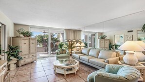 Un salon spacieux avec un grand canapé, une table basse en verre et un coin repas avec des chaises. Il y a un balcon agrémenté de plantes et offrant une vue sur l’extérieur.