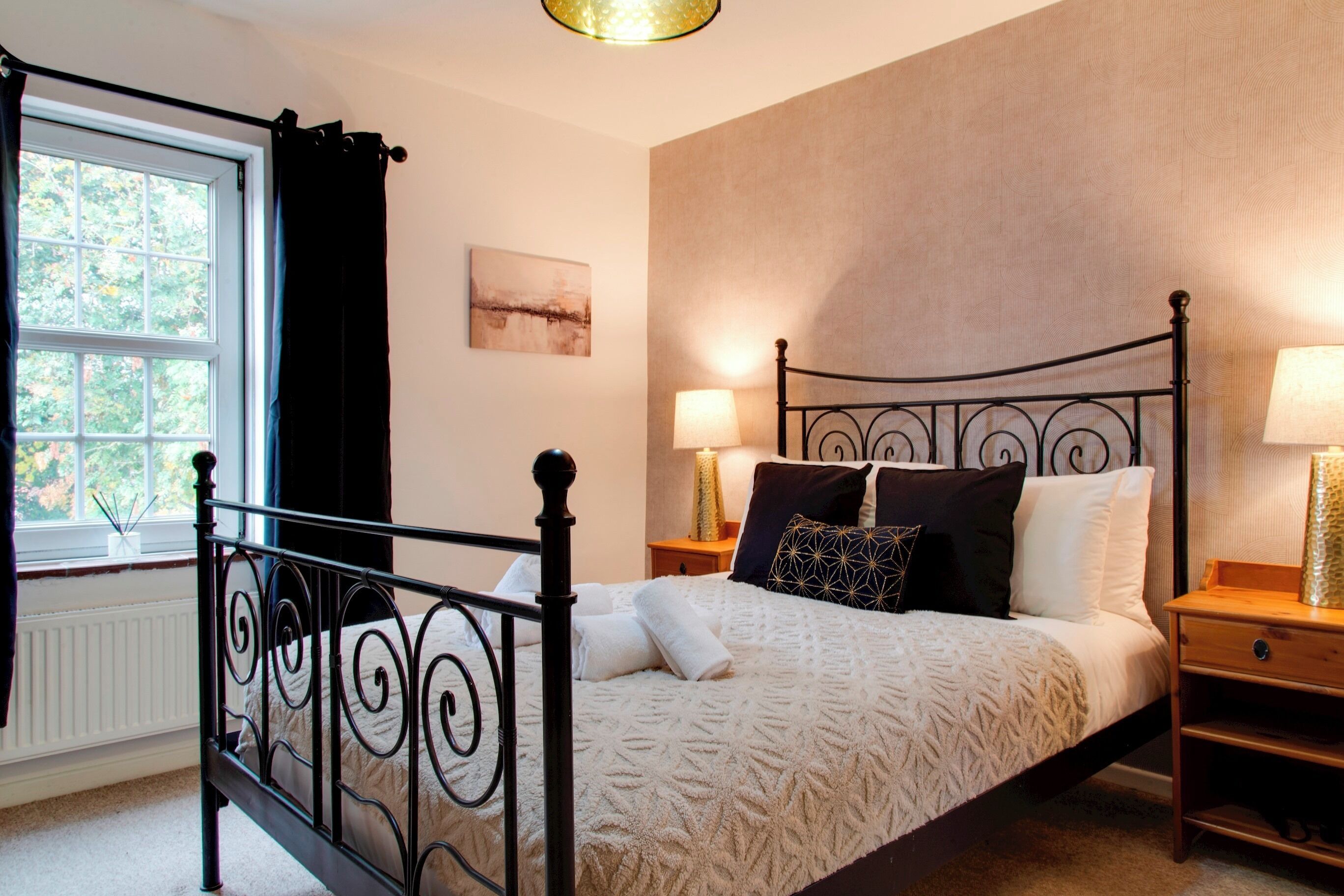 A bedroom with a bed, a window with curtains, a bedside table with a lamp, and a wall-mounted artwork.