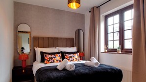 A neatly made bed with a black cover, two pillows with a multicolored pattern, and two rolled towels. There is a round mirror on a bedside table, a red lamp, and a window with curtains. - Stylish Victorian Terrace in Central Cambridge | Pass the Keys (Cambridge)