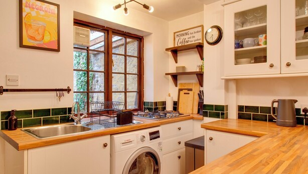 A compact kitchen with a washing machine, a sink, and a window with a view of greenery. - Stylish Victorian Terrace in Central Cambridge | Pass the Keys (Cambridge)