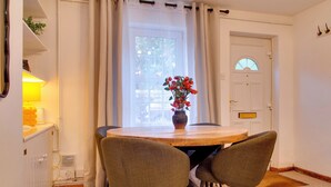 A dining area with a round wooden table, two chairs, a vase with flowers, a window with curtains, a radiator, and a door with a small window. - Stylish Victorian Terrace in Central Cambridge | Pass the Keys (Cambridge)
