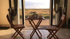 A dining area with a table and chairs, a view of a field through glass doors, and a small rug on the floor. - Pass the Keys | Levante Coastal Cabin - Dungeness, Sleeps 2/3 (Folkestone)