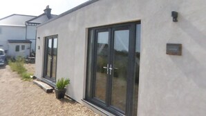 A modern building with large sliding glass doors, a small outdoor area with a potted plant, and a gravel driveway. - Pass the Keys | Levante Coastal Cabin - Dungeness, Sleeps 2/3 (Folkestone)