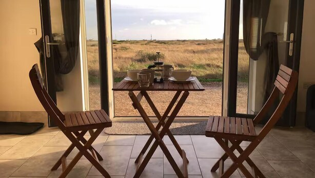 A dining area with a table and chairs, a view of a field through glass doors, and a small rug on the floor. - Mistral Coastal Cabin - Dungeness, sleeps 2/3 - Pass the Keys (Folkestone)