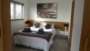 A bedroom with a bed, a nightstand, a lamp, a window with blinds, and a painting on the wall. - Mistral Coastal Cabin - Dungeness, sleeps 2/3 - Pass the Keys (Folkestone)