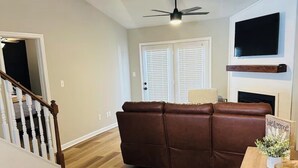 A modern living room with a brown leather sofa, wooden flooring, a ceiling fan, and a flat-screen TV mounted above a fireplace.