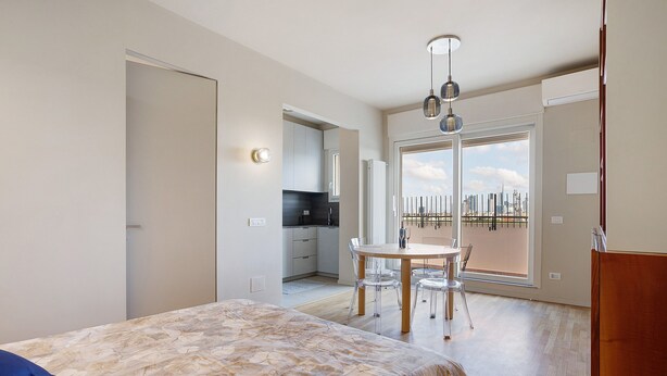 Ein Schlafzimmer mit Bett, Esstisch mit Stühlen, Küchenzeile und einem großen Fenster mit Stadtblick.